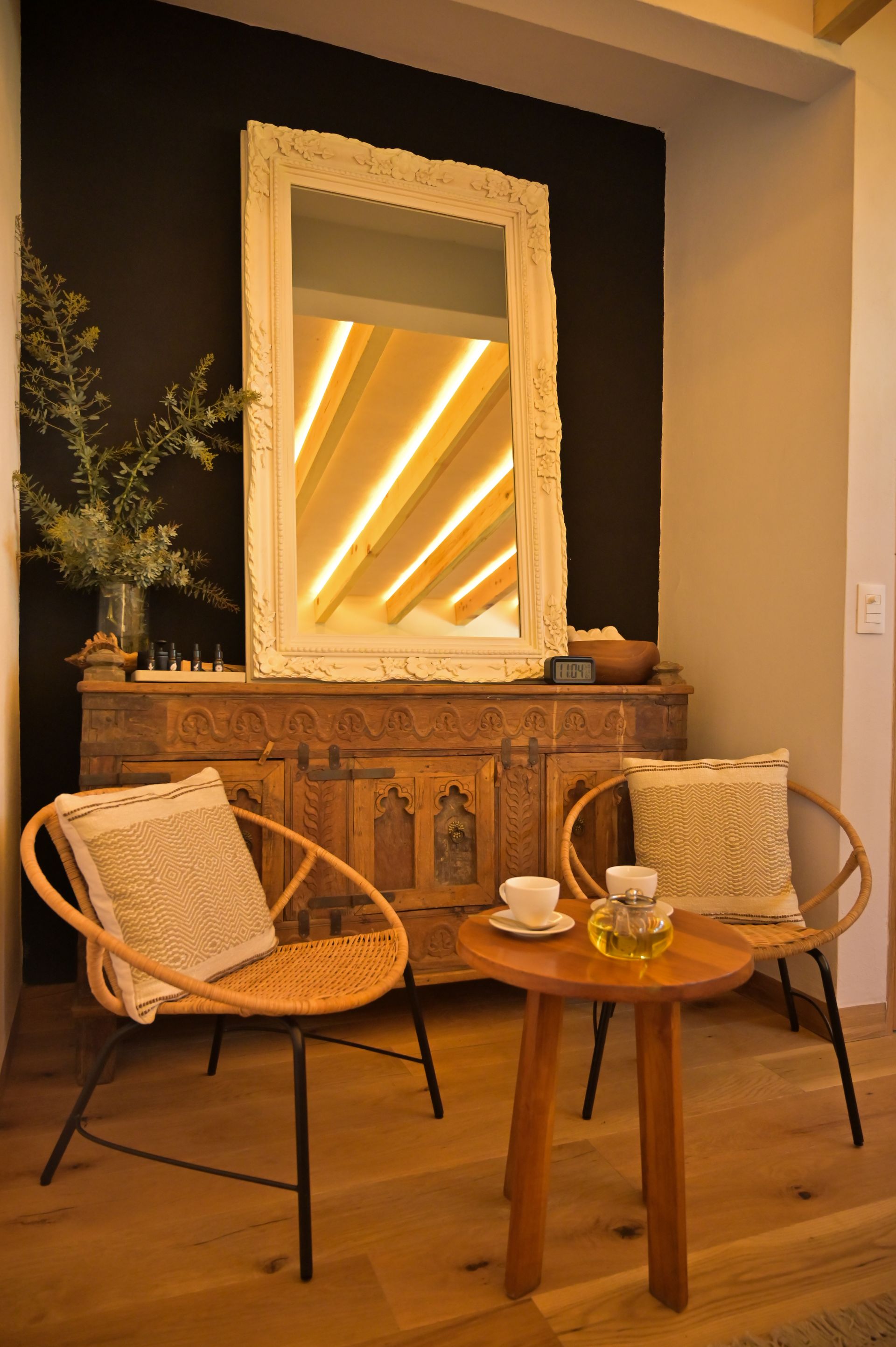 Acogedor salón con sillas de mimbre, mesa redonda de madera, espejo ornamentado e iluminación ambiental cálida.