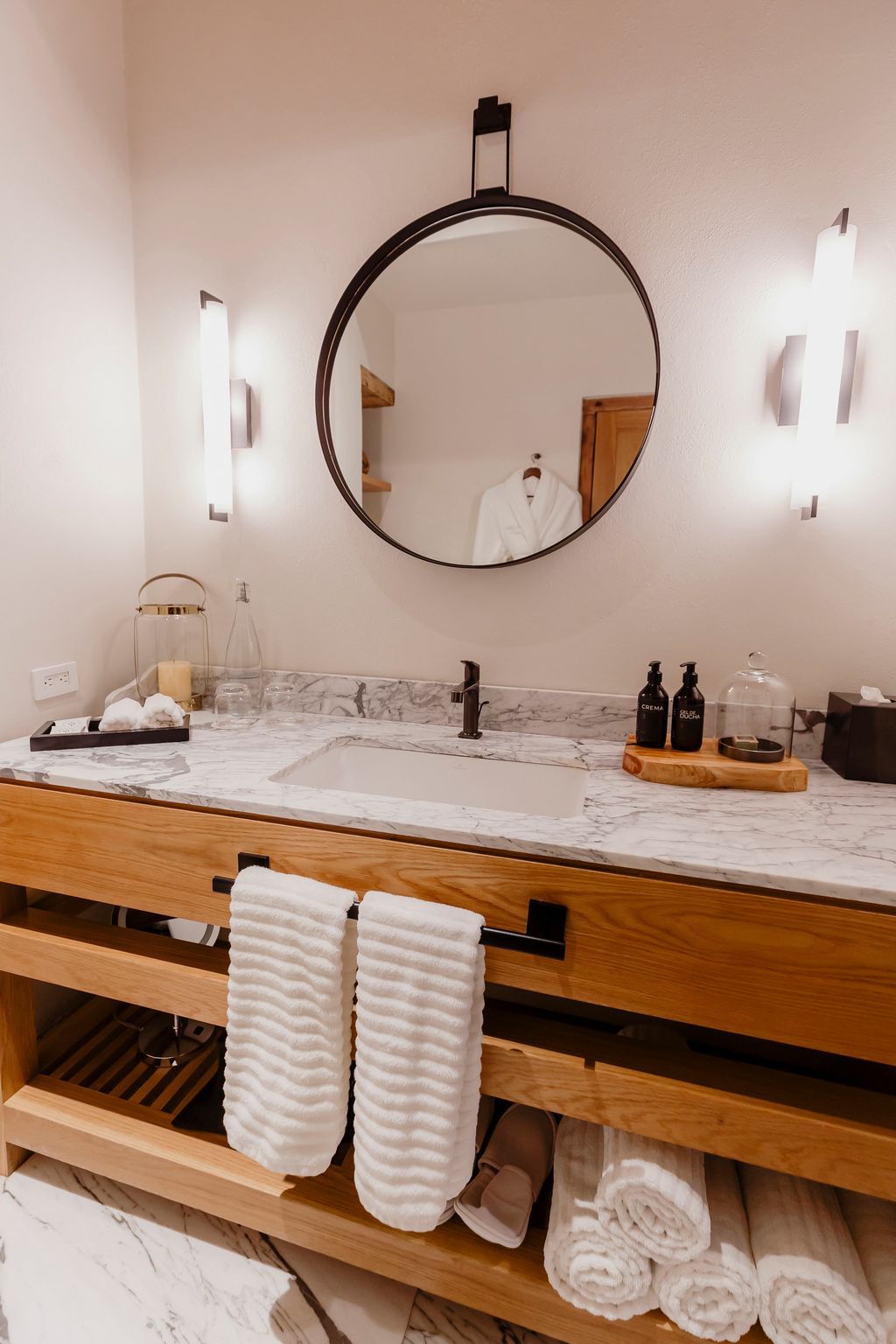 Tocador de baño moderno con espejo redondo, encimera de mármol, lavabos y toallas blancas cuidadosamente apiladas.