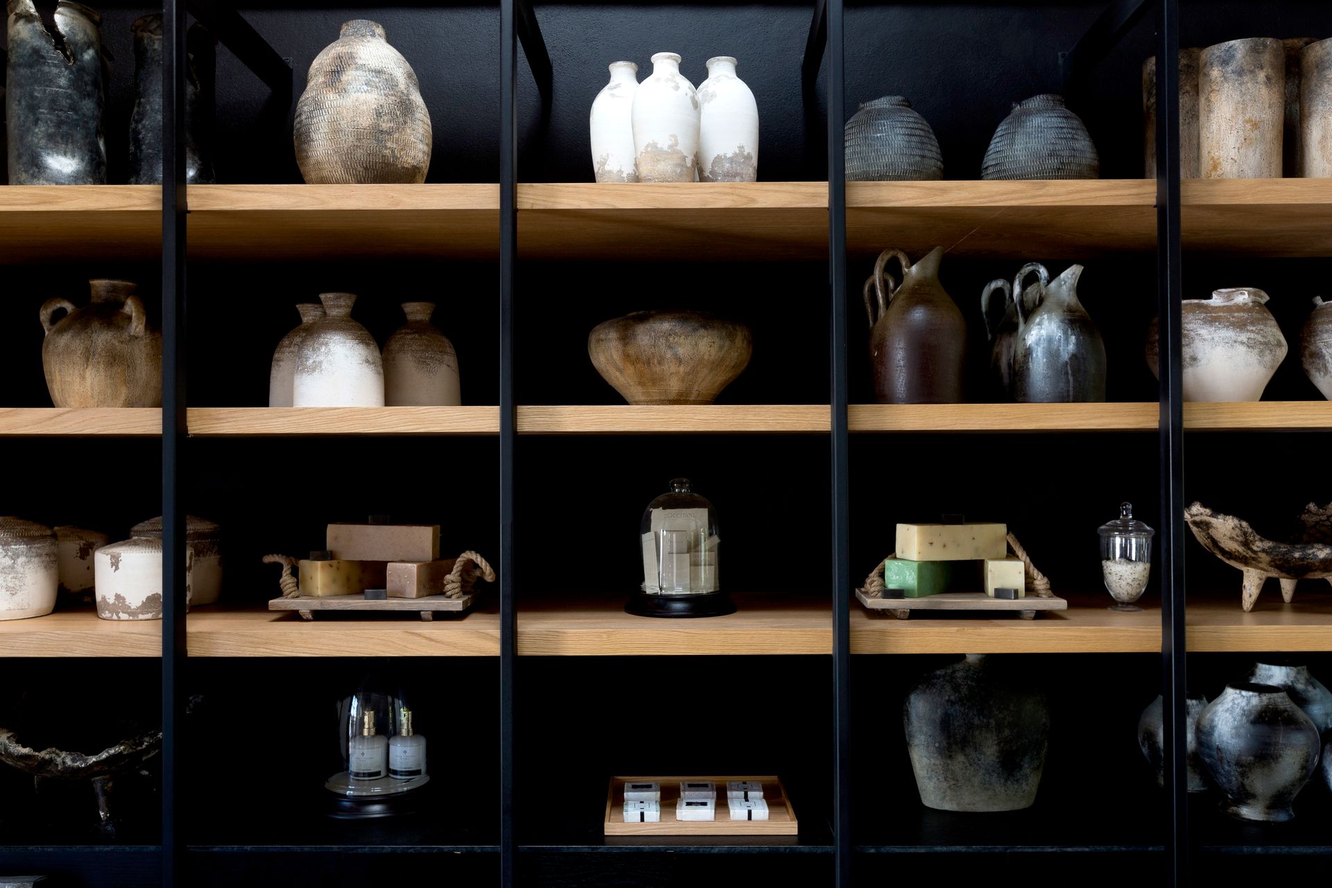 Exhibición en estantes de cerámica variada, botellas y objetos pequeños sobre una pared de rejilla oscura.