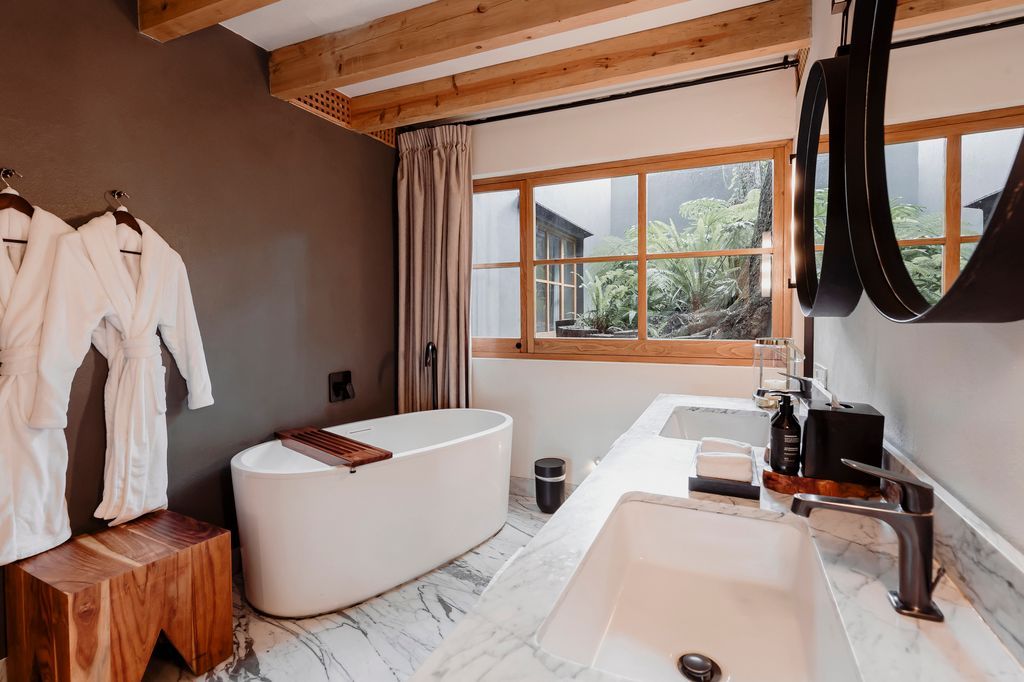 Baño moderno con detalles en madera, bañera independiente, lavabos dobles y una gran ventana con vistas a la vegetación.