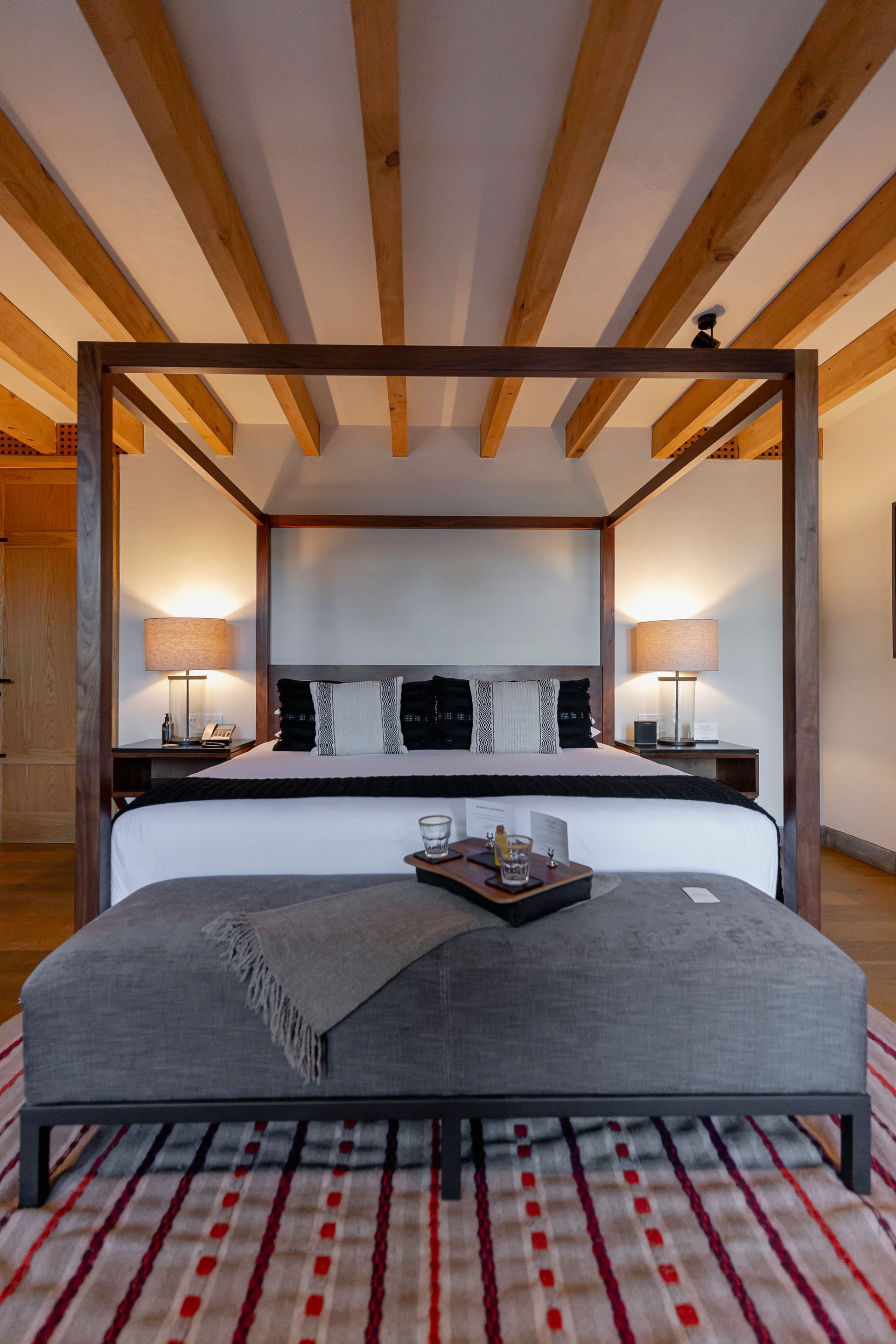 Dormitorio moderno con cama con dosel, vigas de madera a la vista, banco gris e iluminación tenue con lámparas.
