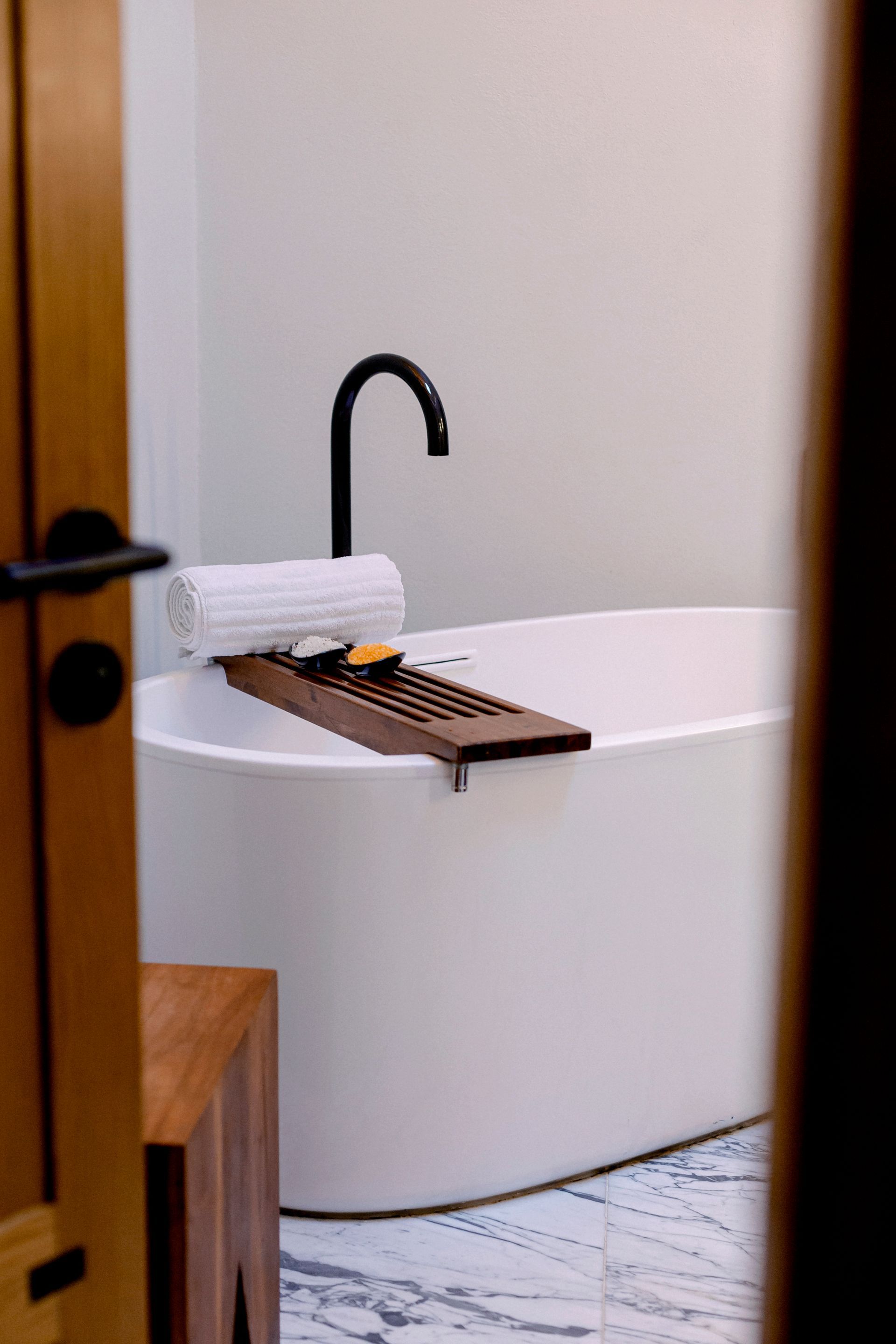 Bañera blanca independiente con grifo negro, bandeja de madera y toalla enrollada en un baño.