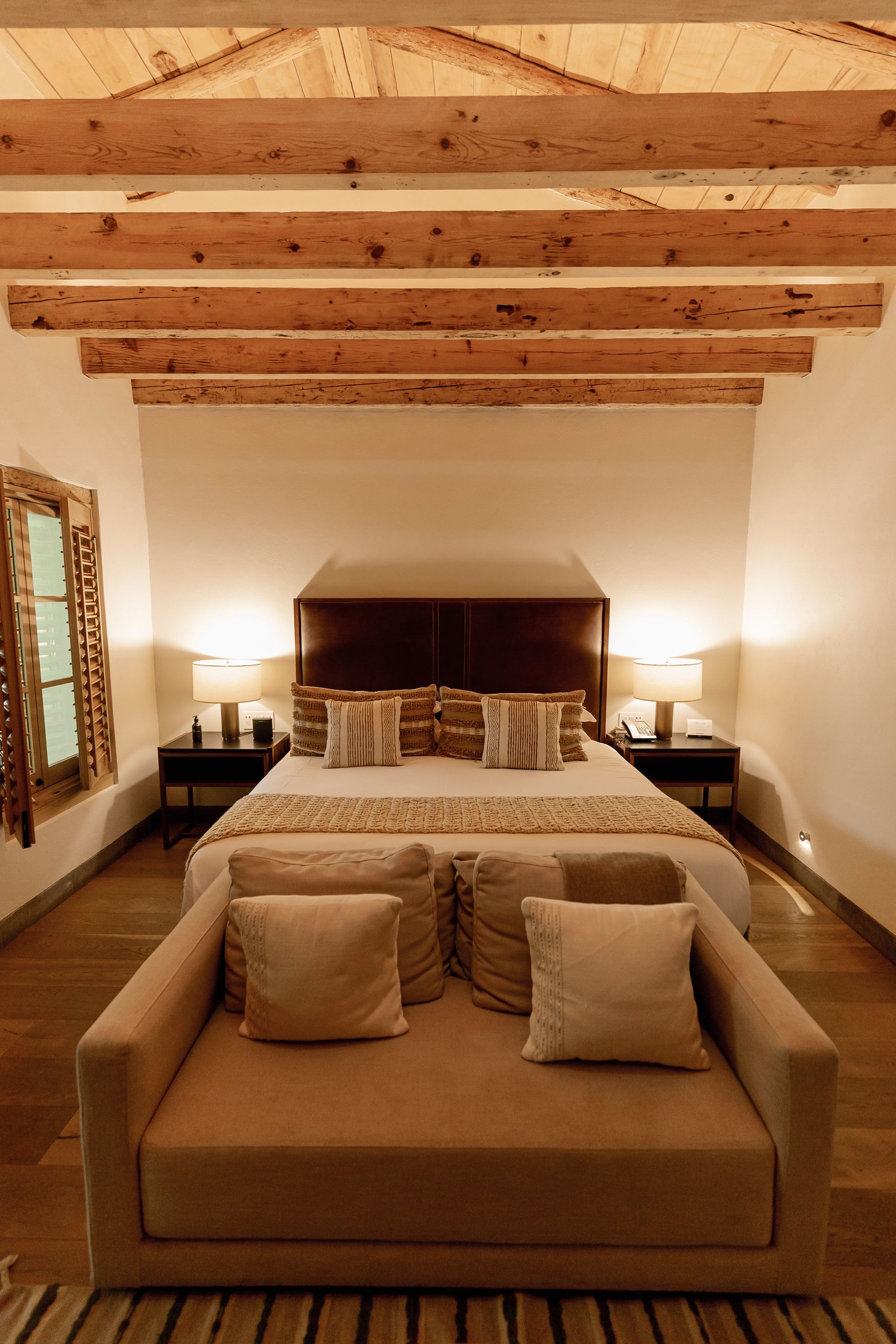 Habitación acogedora con techo de vigas de madera, cama grande y salita privada con ofá beige a los pies de la cama.
