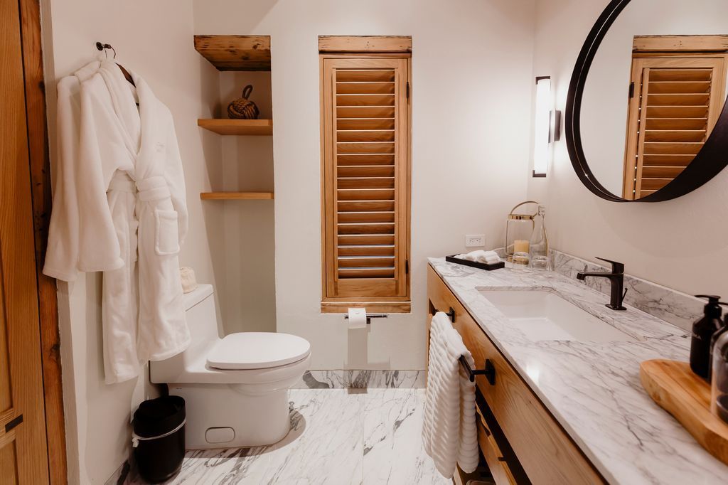 Baño moderno con albornoces blancos, tocador de madera, espejo redondo, inodoro, ducha y detalles de mármol.