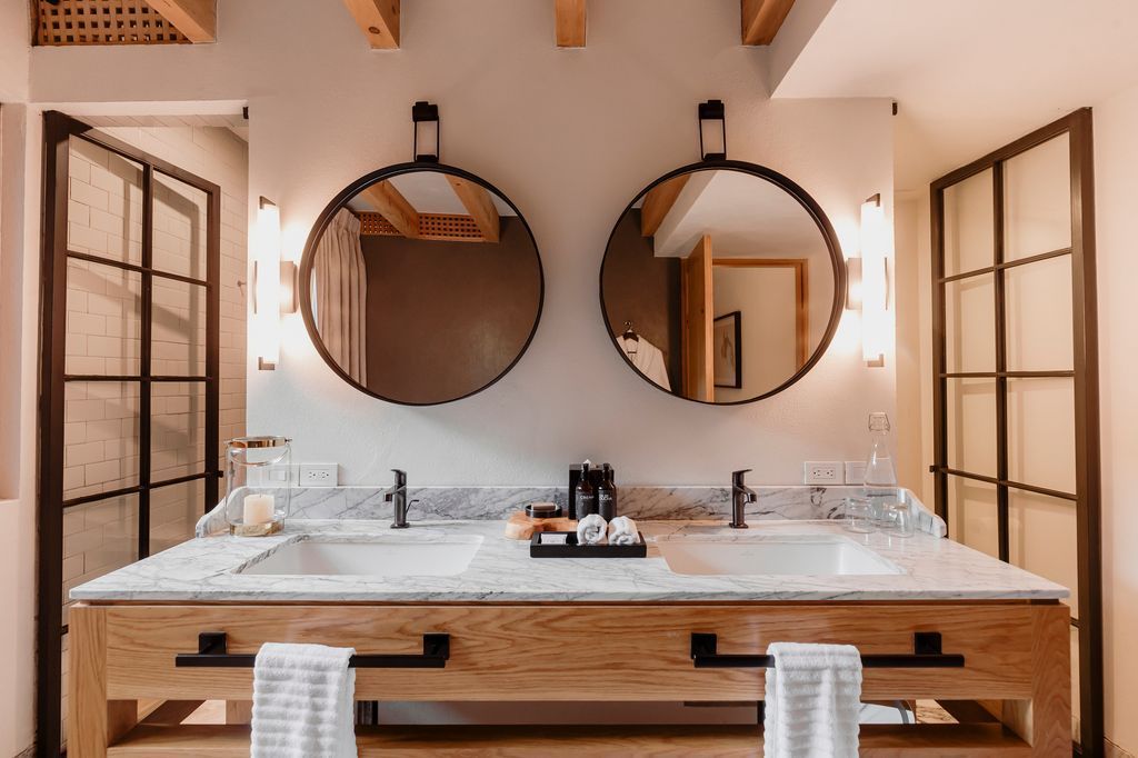 Tocador de baño moderno con dos espejos redondos, encimera de mármol y detalles de madera.