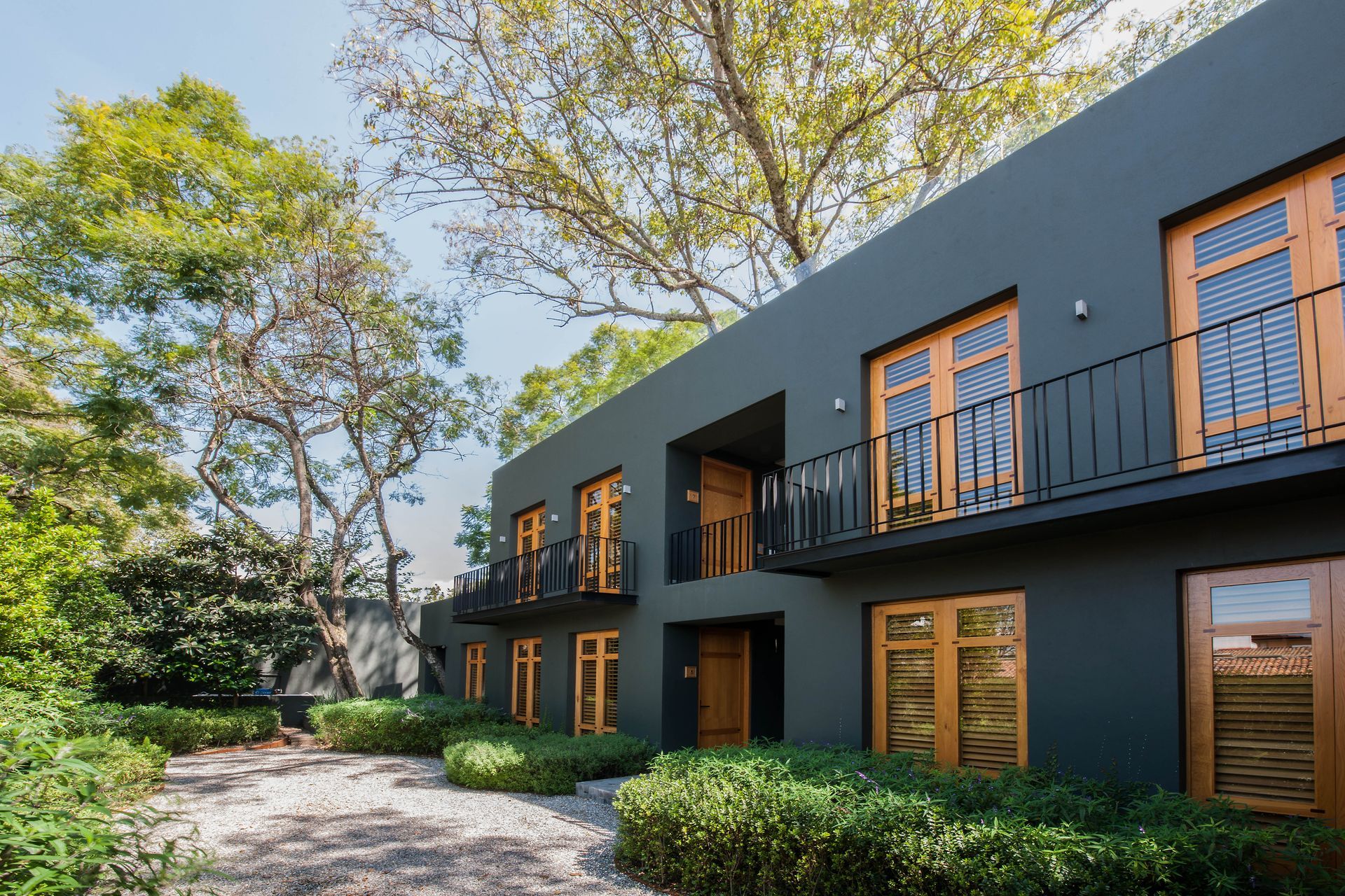 Edificio de apartamentos moderno de color gris oscuro con puertas y balcones de madera, junto a árboles y un camino de grava.