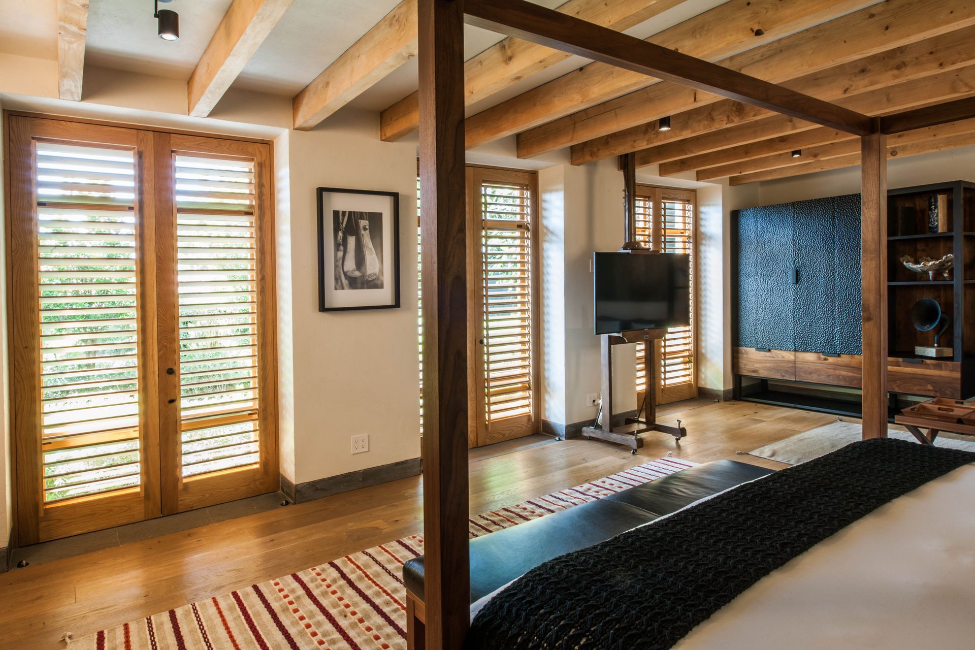 Dormitorio moderno con vigas vistas, contraventanas de madera, una cama y un televisor montado en la pared.