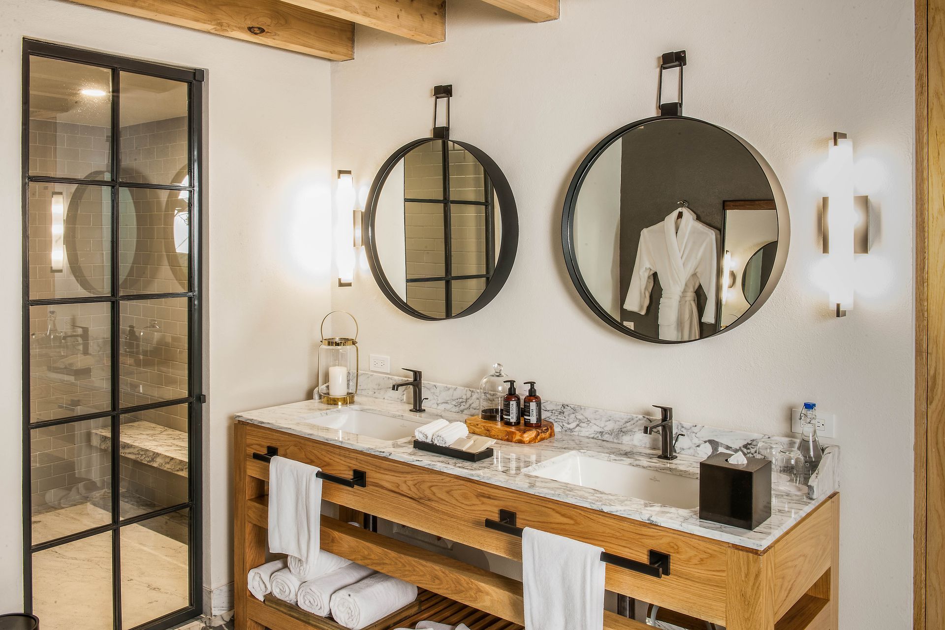 Baño moderno con dos espejos redondos, tocador de madera y puerta de ducha de cristal.