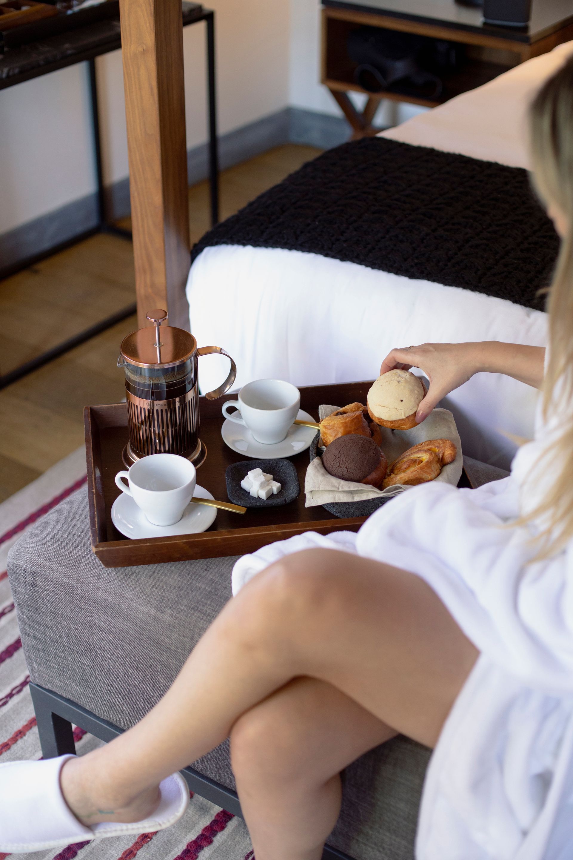 Bandeja de desayuno sobre la cama con té, pasteles y una persona sentada con una bata blanca.