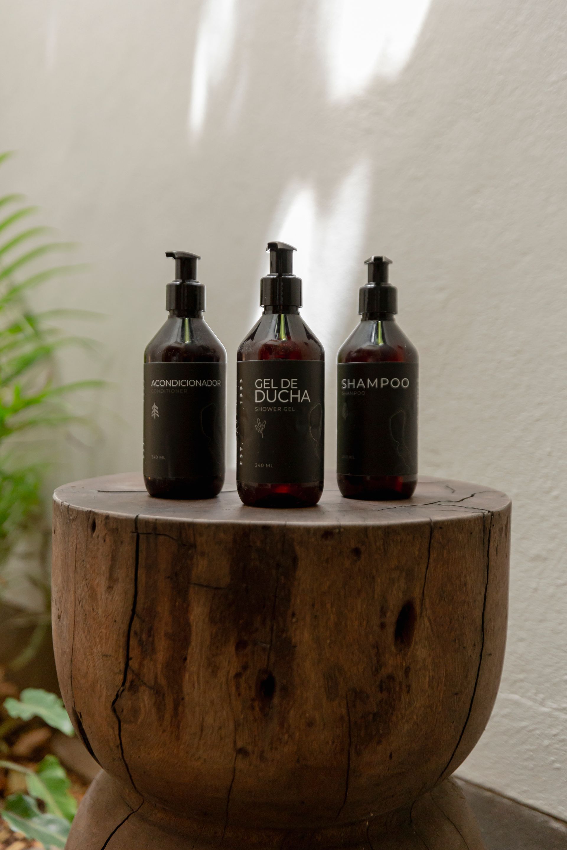 Tres botellas oscuras con dosificador sobre un tocón de madera contra una pared clara con sombras suaves.