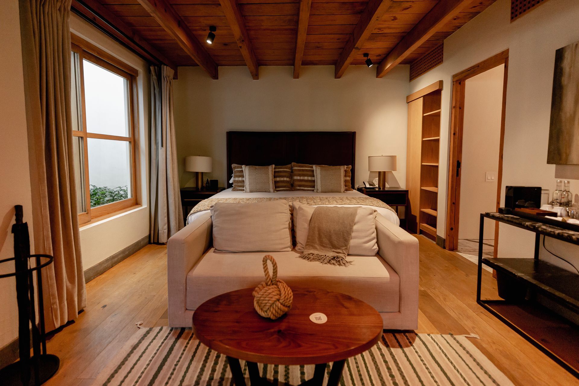 Acogedora habitación con techo de madera, cama grande, sofá y cálida luz natural que entra por las ventanas.