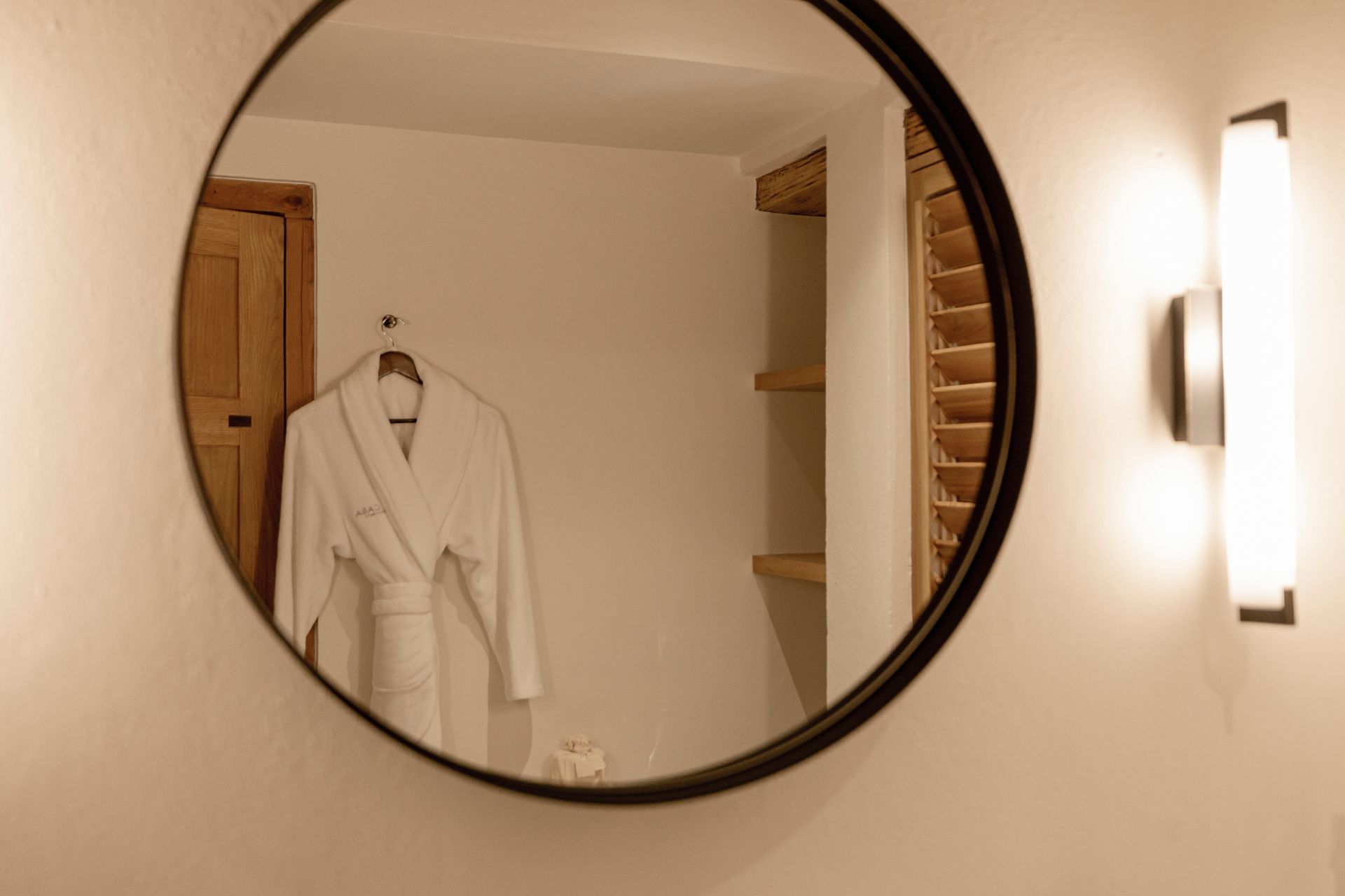 Espejo de pared redondo que refleja una bata blanca en una habitación con iluminación cálida, estantes y un aplique de pared.