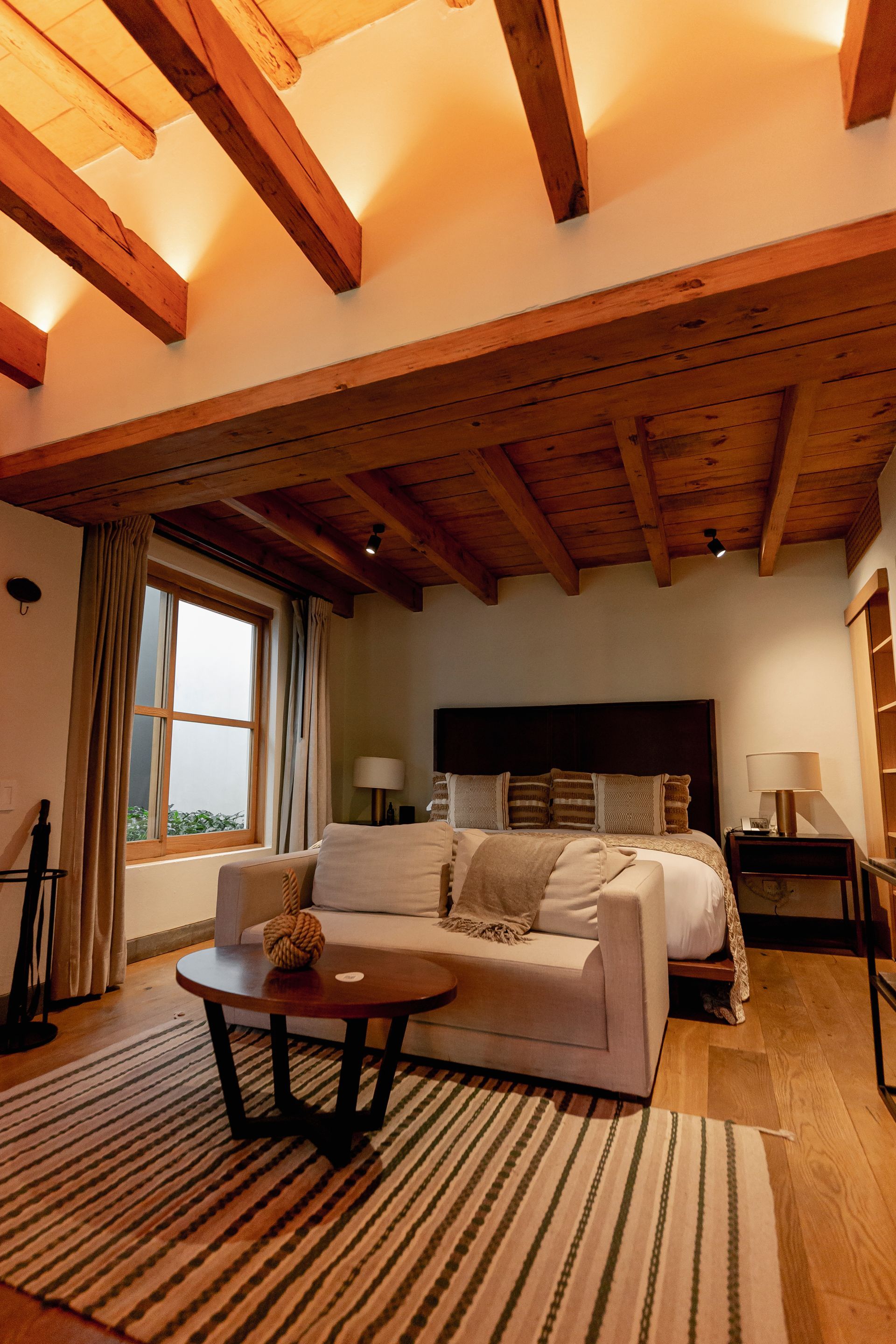 Acogedora sala de estar con vigas de madera, sofá beige, alfombra a rayas y una ventana con vistas a la montaña.