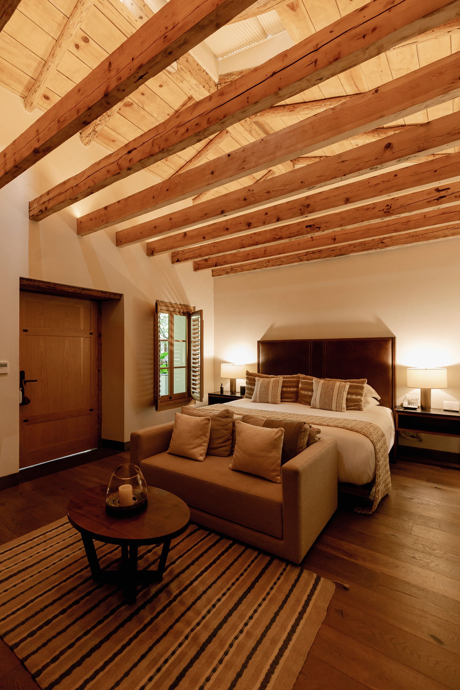 Acogedora habitación con vigas de madera a la vista, una cama grande, sofá e iluminación cálida sobre una alfombra a rayas.