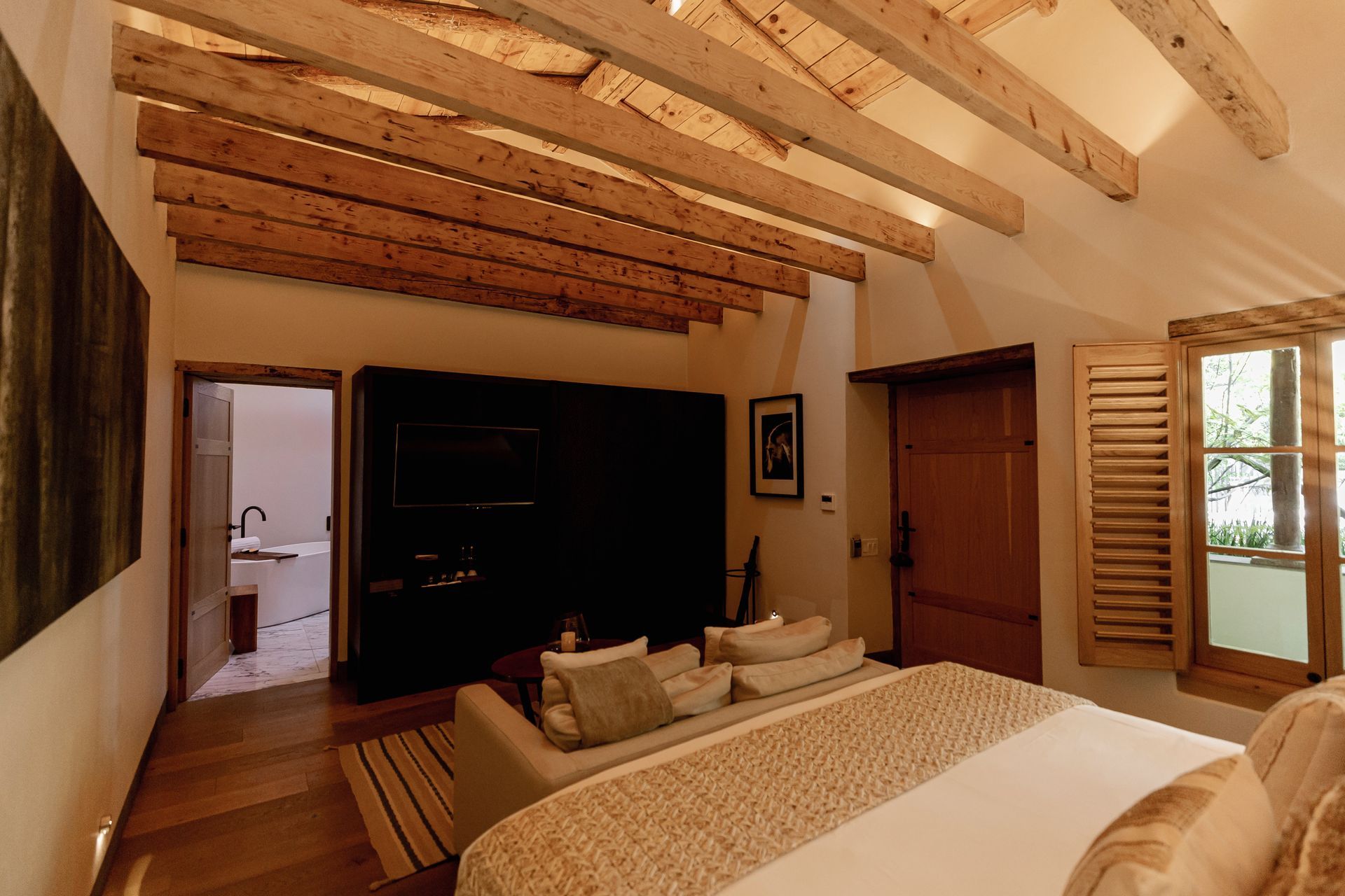 Acogedora habitación con techo de vigas de madera, cama king size, sofá, armario oscuro y ventana con vistas a la vegetación.