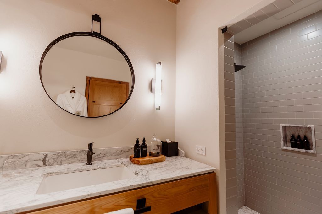 Mueble de baño con espejo redondo, encimera de mármol y cabina de ducha con azulejos estampados.