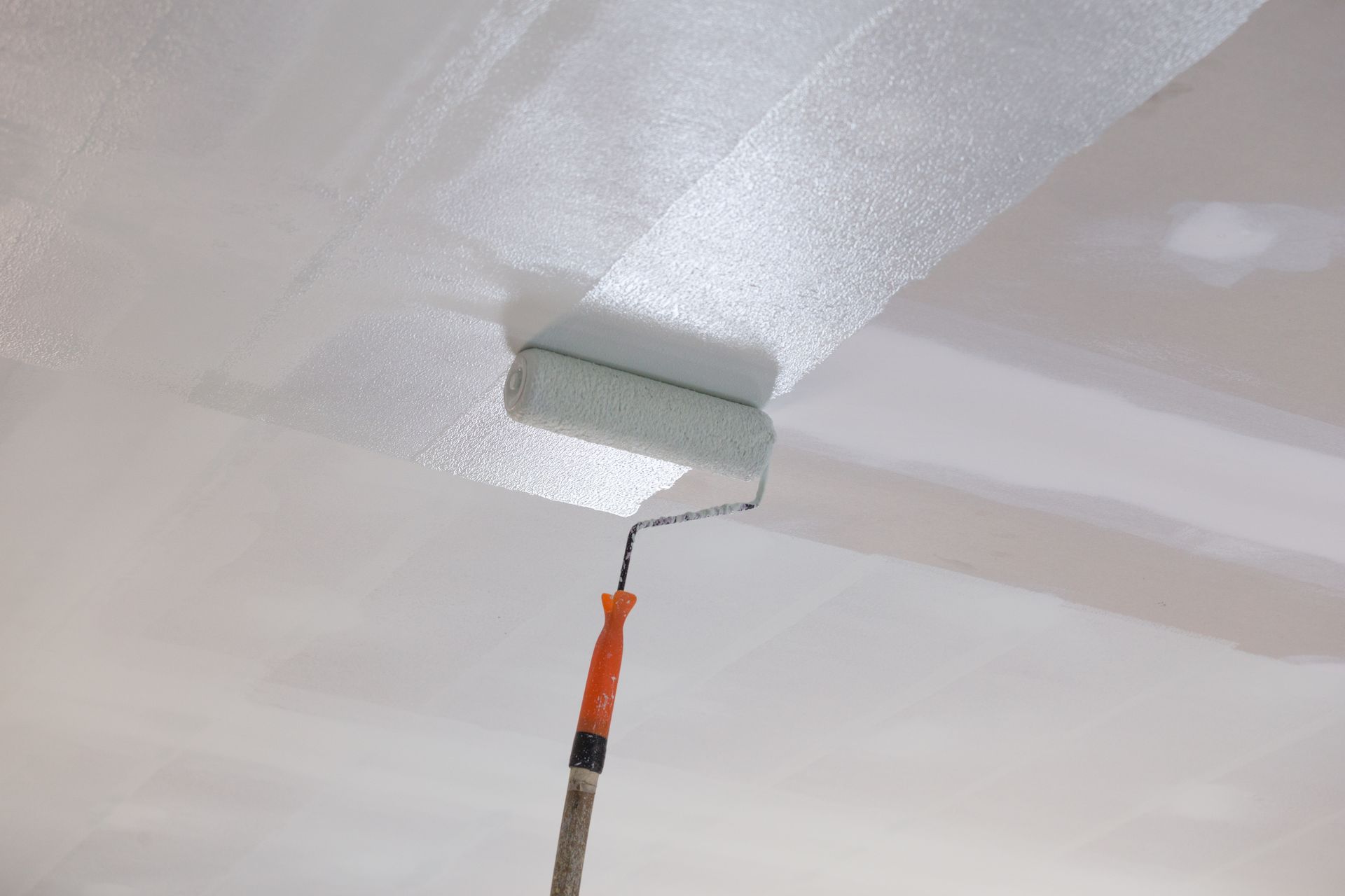Paint roller applying white paint to a ceiling, covering a patch near a corner.