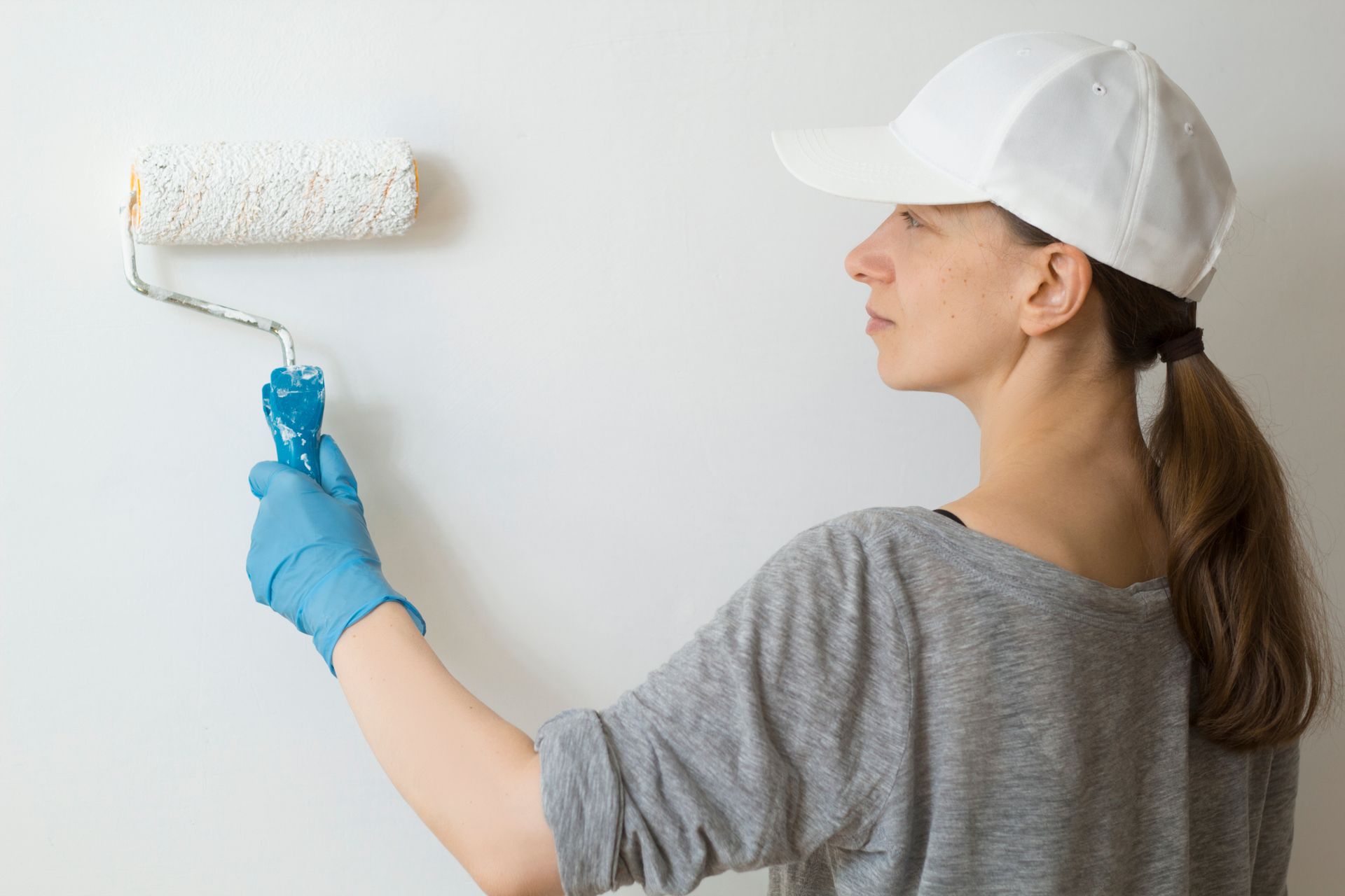 Person in a white cap and blue gloves painting a white wall with a roller.