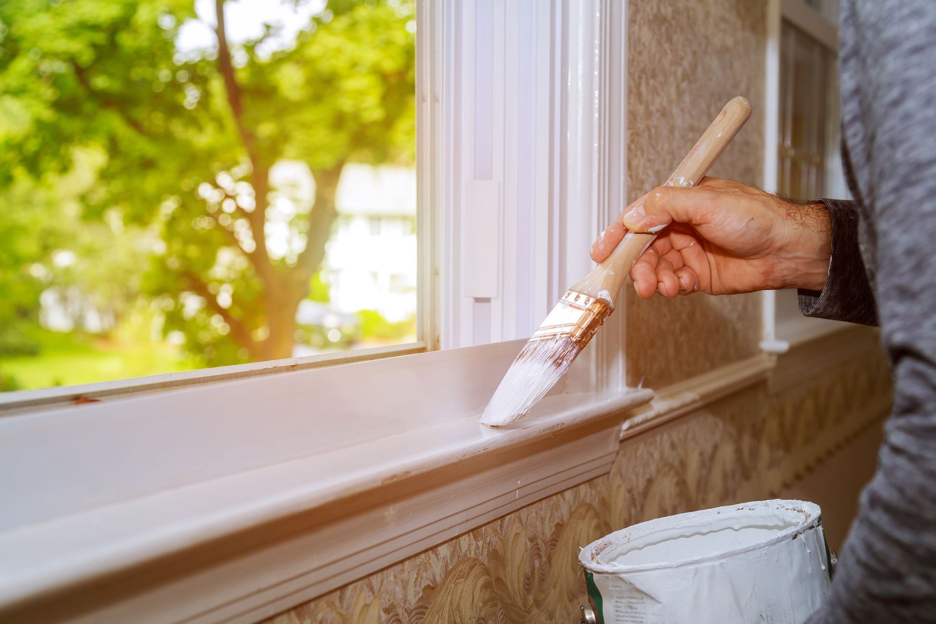 Hand painting white trim along a window frame with a brush, paint bucket below.