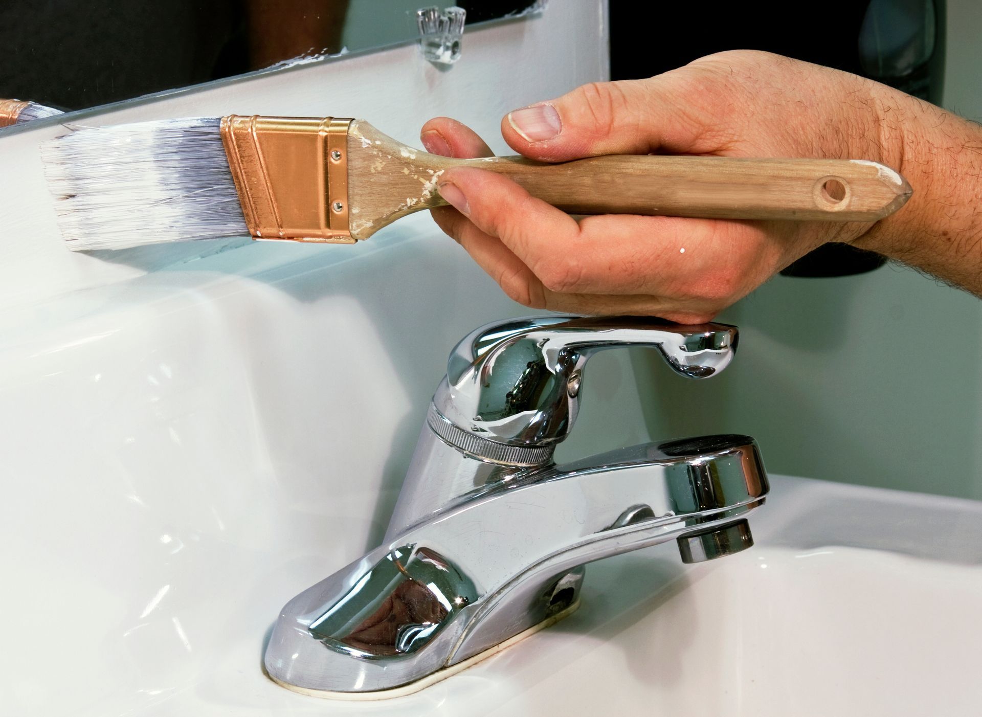 A hand using a paintbrush to paint a wall above a bathroom sink faucet