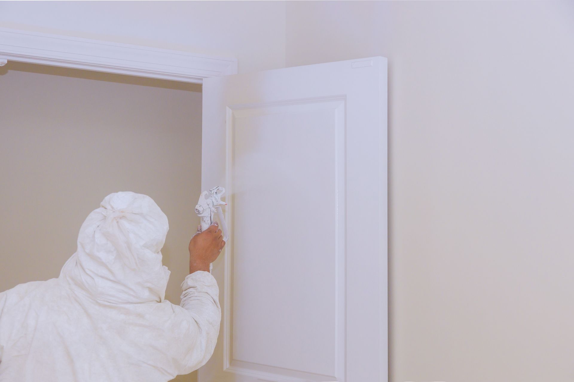 Person in white coveralls painting a white door frame in a beige room.