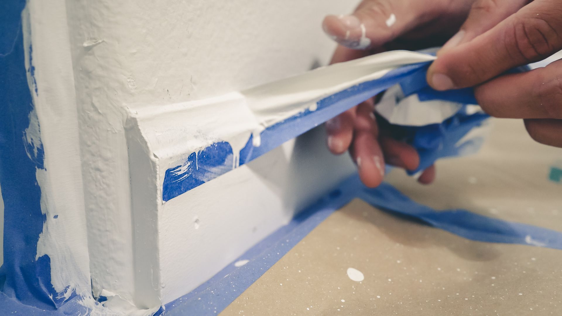 Hand peeling blue painter’s tape from a white baseboard during painting prep