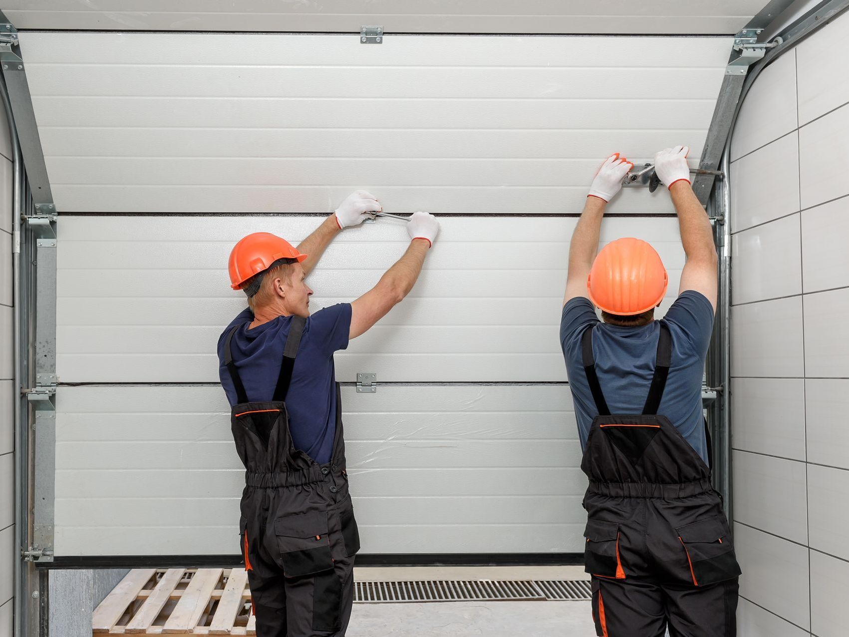 Deux ouvriers portant des casques et des gants orange installent une porte de garage blanche.