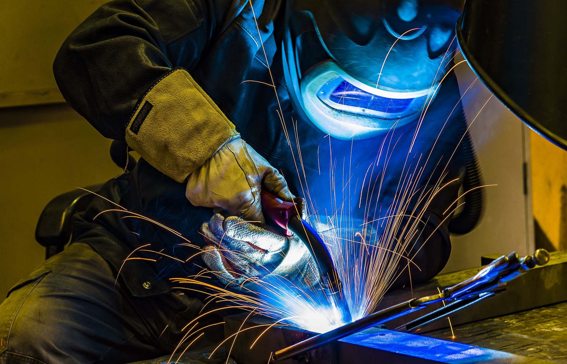 Soudeur dans un atelier portant un équipement de protection, soudant du métal avec une lumière bleue vive et des étincelles.