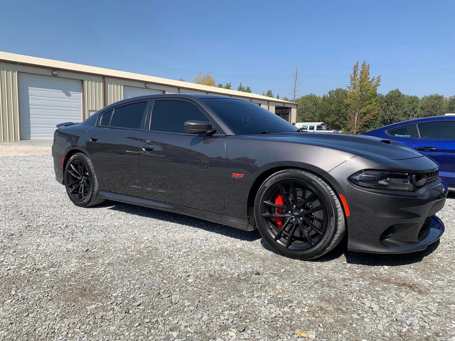 Dark gray Dodge Charger, black rims, parked in a gravel lot on a sunny day. Red brake calipers visible.
