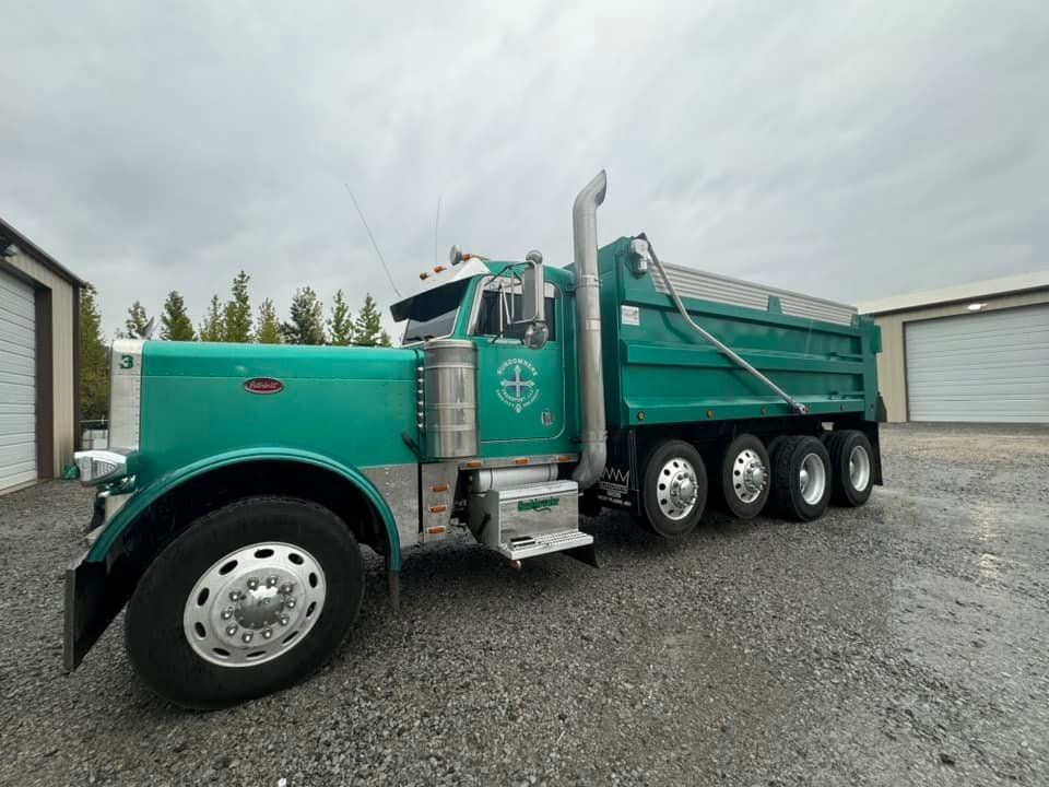 Green Peterbilt dump truck parked in front of a gray garage under overcast sky.