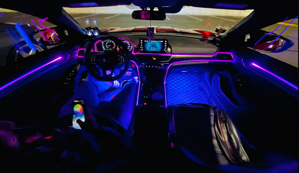 Car interior with purple LED lights. Person adjusts lights with a phone, driving on a road.
