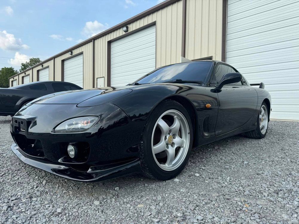 Black Mazda RX-7 sports car parked in front of a building with garage doors, under a partly cloudy sky.