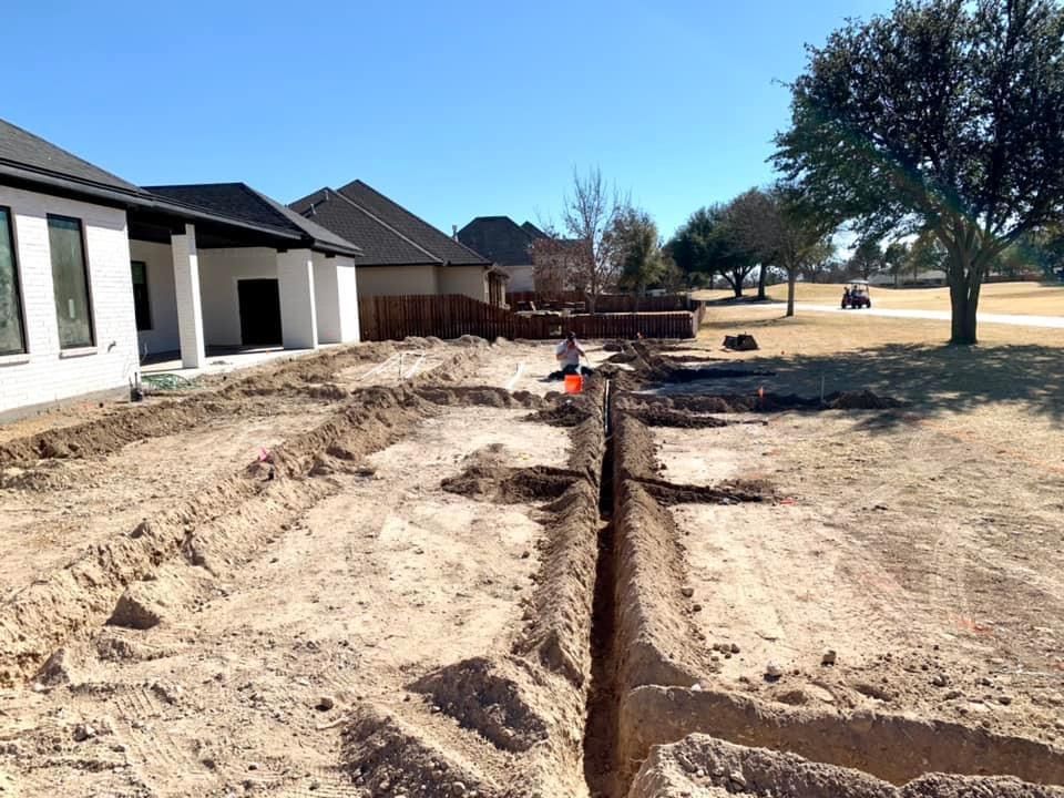 Trenches dug in yard next to houses, a person works in the distance; sunny day, golf course view.