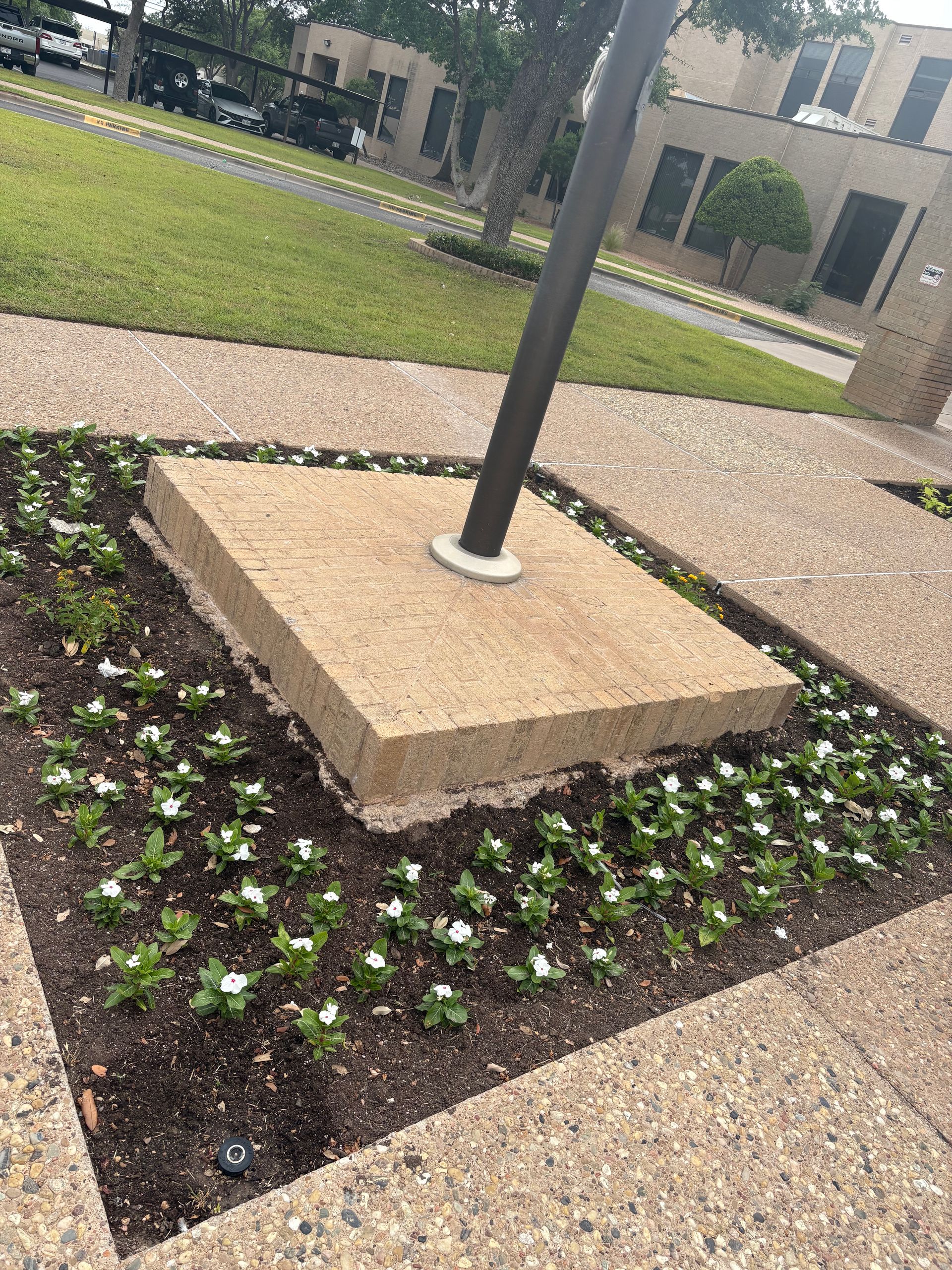 Concrete base with flagpole in flower bed, surrounded by a paved walkway and green grass.