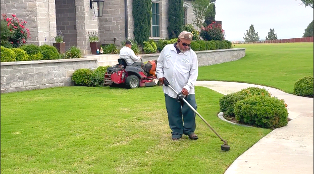 Man using weed whacker on lawn's edge, another man mowing in the background, large house.