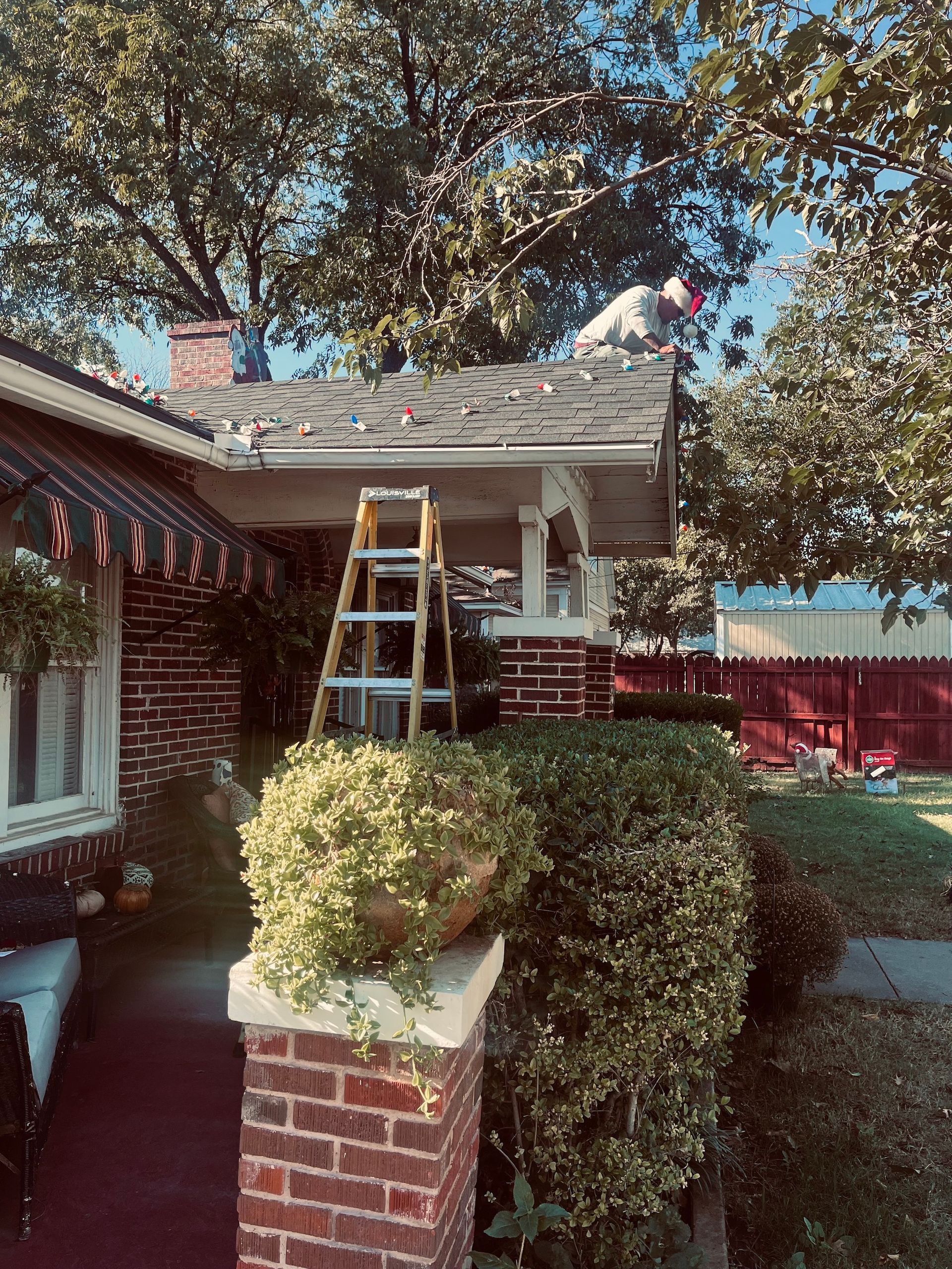 Person on a ladder fixing roof; brick house with porch, shrubs, and trees.