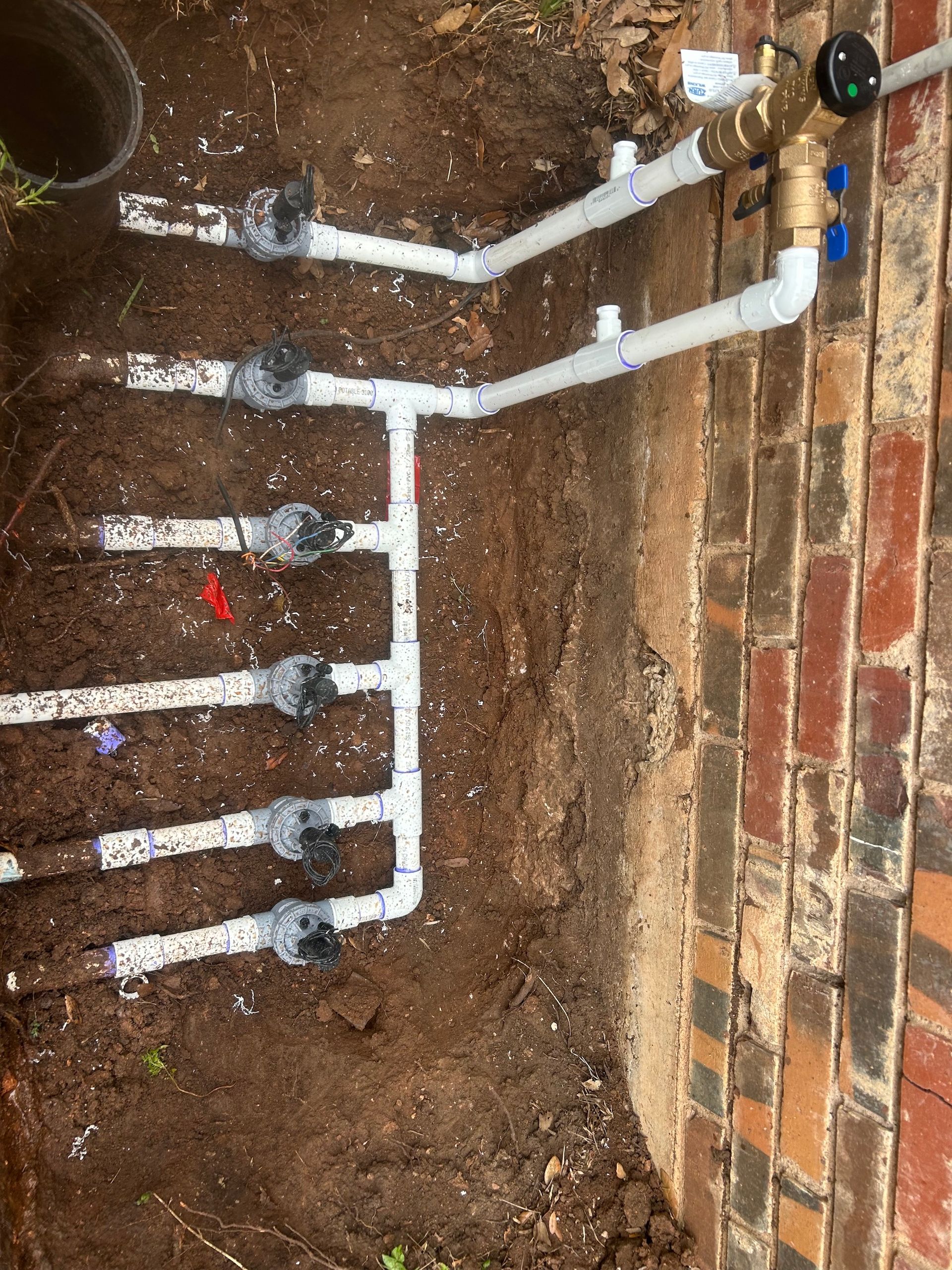 Irrigation system with white PVC pipes, valves, and a brass backflow preventer next to a brick wall.