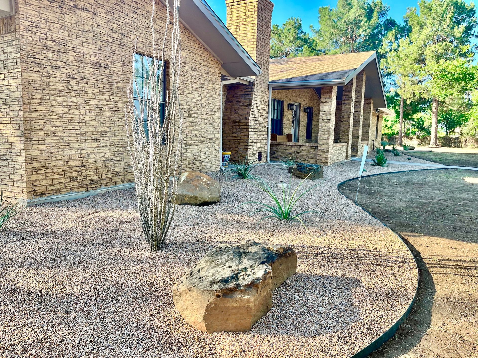 A stone-clad house with a landscaped yard featuring boulders, gravel, and a small plant, sunny day.