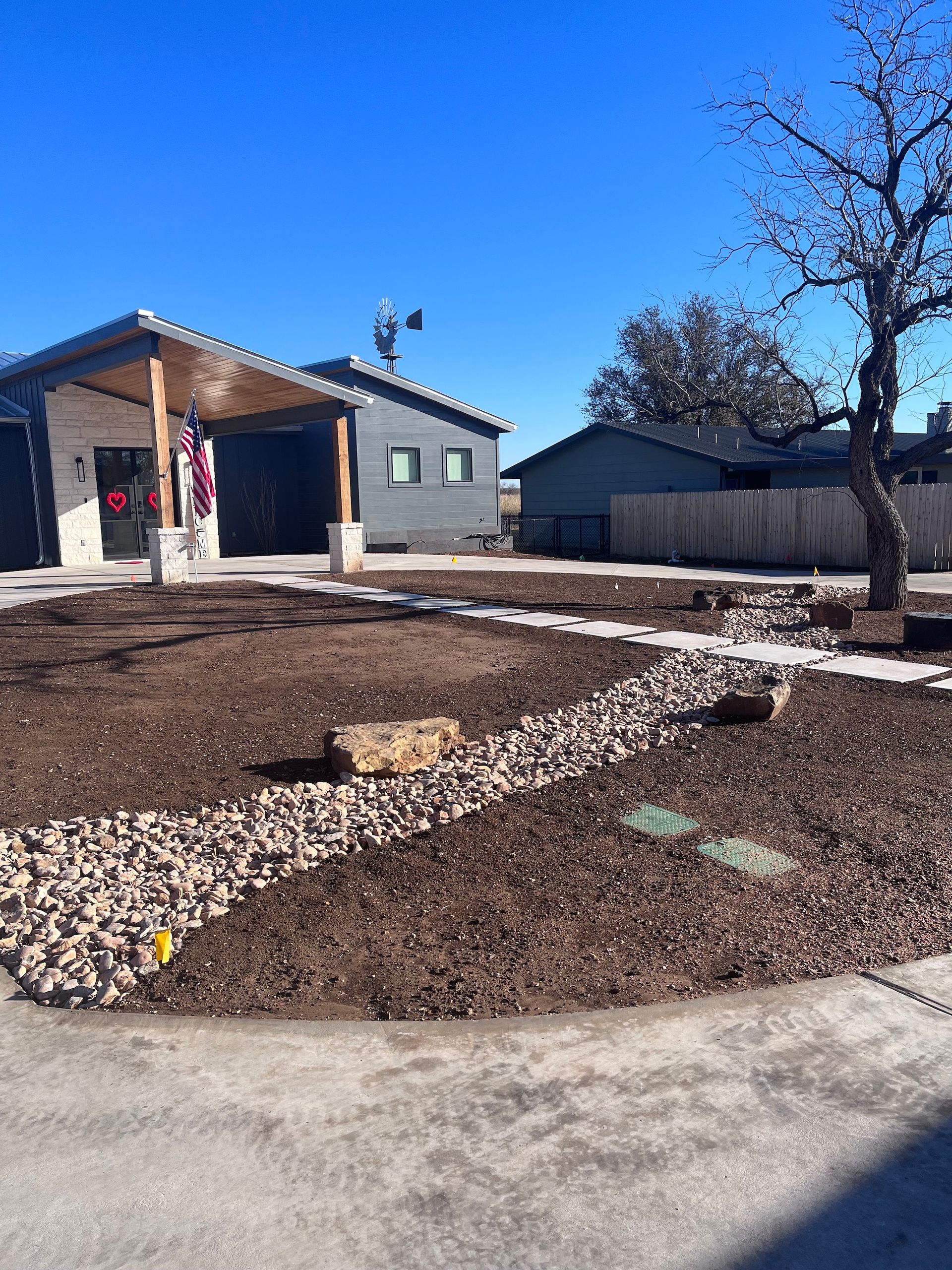 A building with a porch, brown dirt, rocks, and a tree on a sunny day.