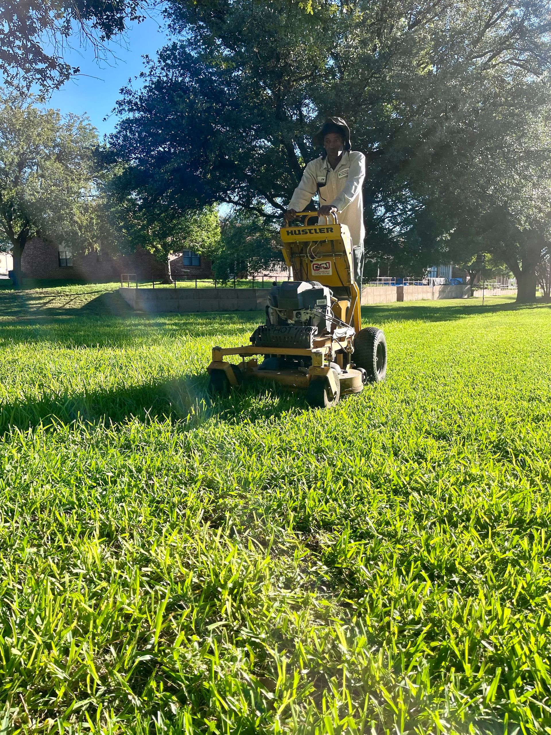 Person mowing a grassy lawn with a large, yellow commercial mower in a park on a sunny day.