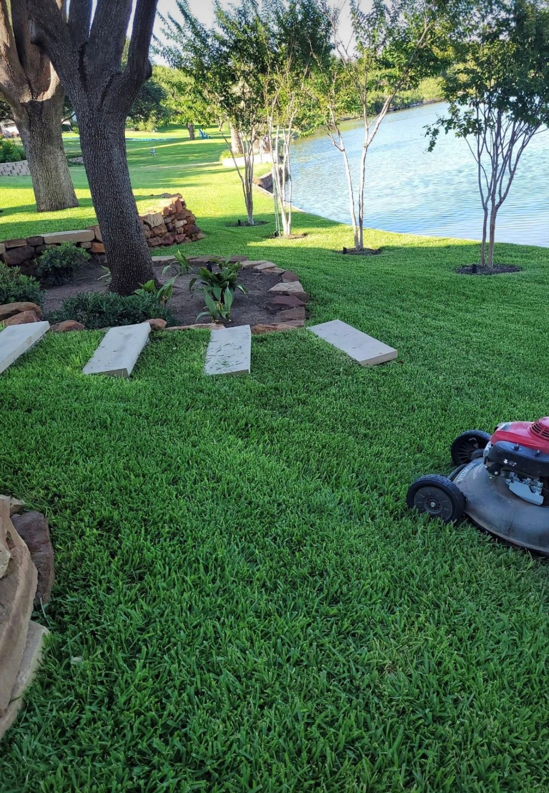 Lawnmower cutting grass near a waterfront. Green lawn, stepping stones, trees, and water.