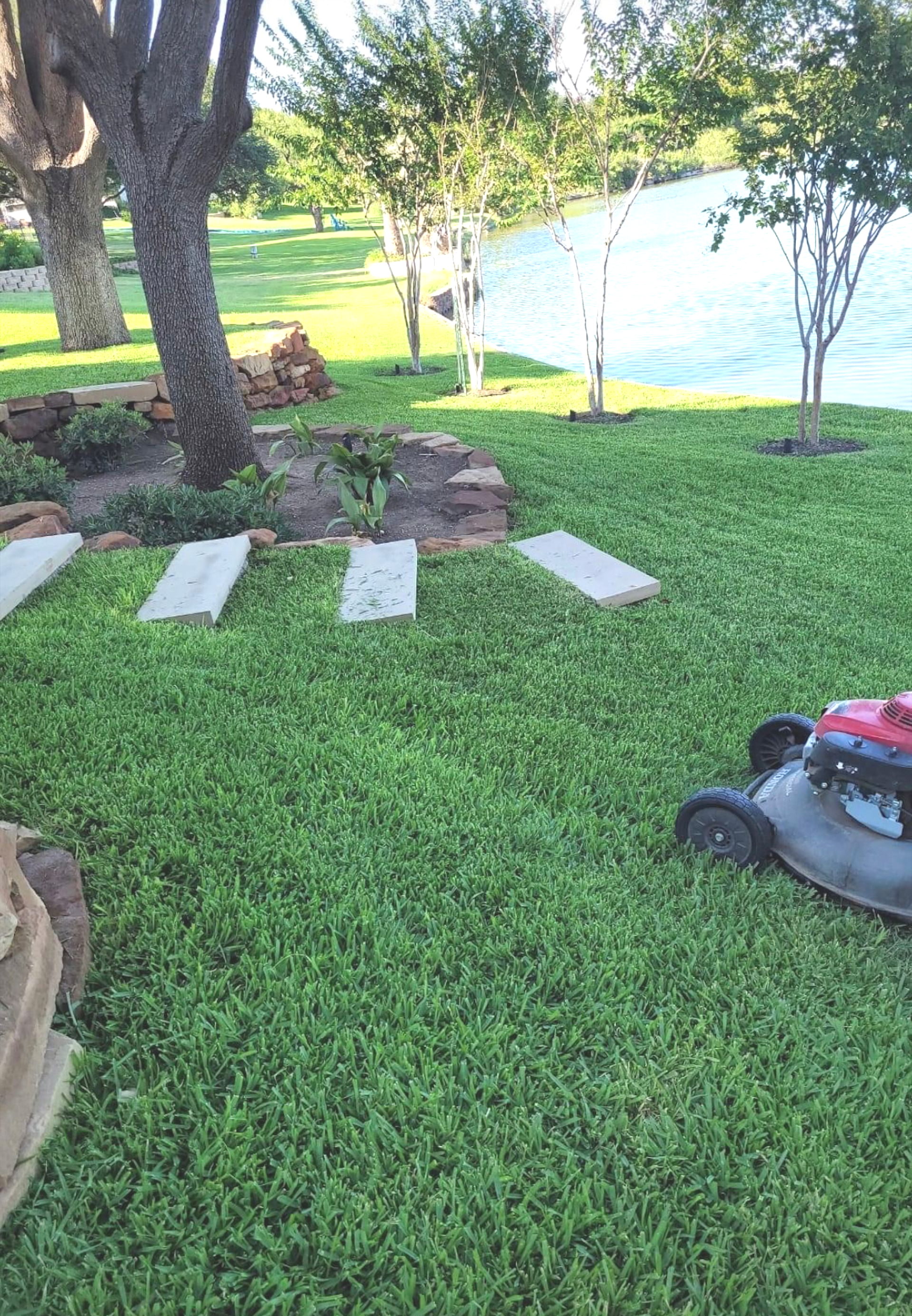 Lawnmower cutting grass near a waterfront. Green lawn, stepping stones, trees, and water.