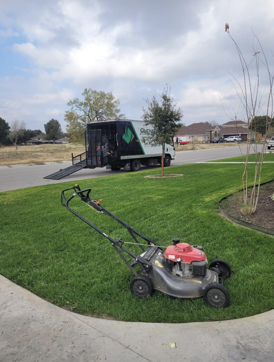Lawnmower on green grass, with a landscaping truck in the background under a cloudy sky.
