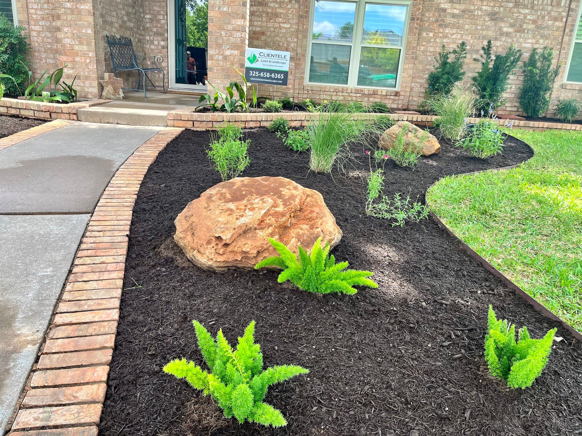 Front yard with a landscaped garden, dark mulch, brick edging, rock, and green plants.