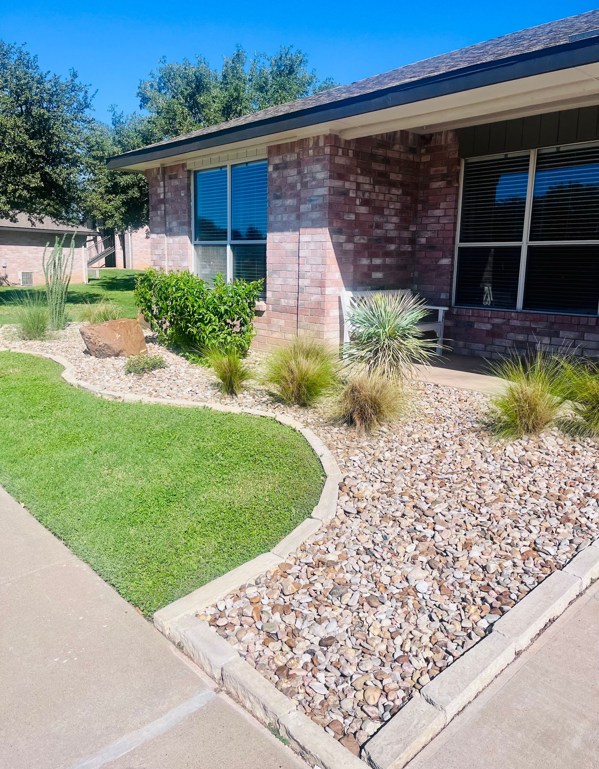 Brick house with landscaped yard, curb separating grass from rock bed with various plants.