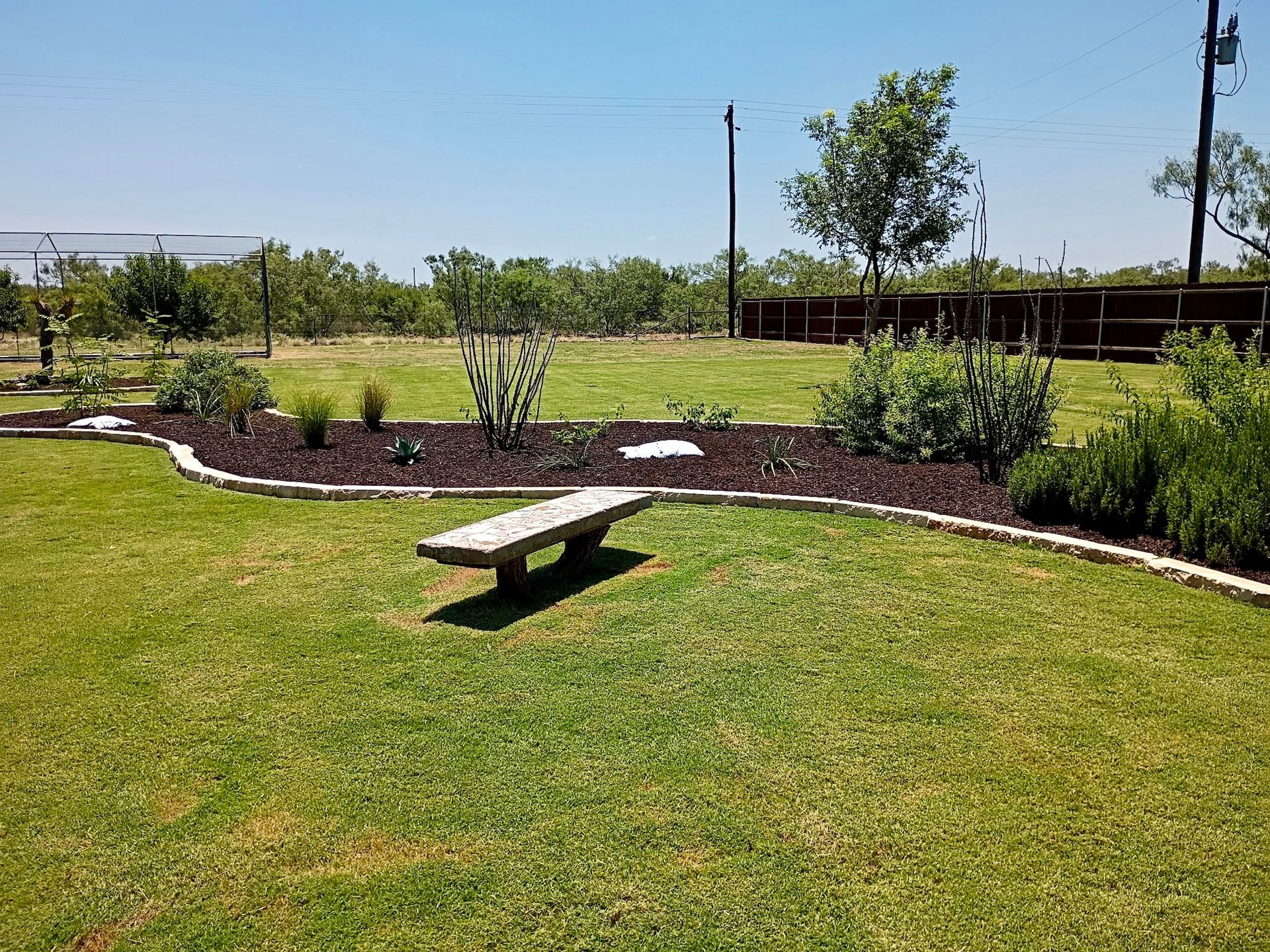 A stone bench sits in a grassy yard bordered by a mulch bed with plants, under a bright blue sky.
