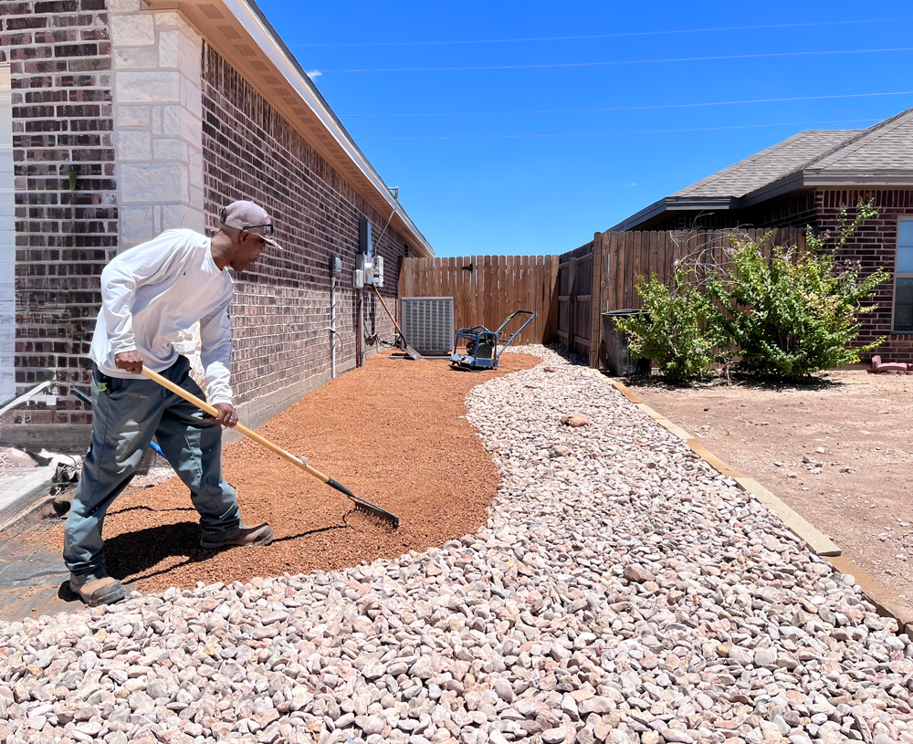 Man rakes mulch into a pathway, bordered by tan rocks, next to a brick building and a wooden fence.