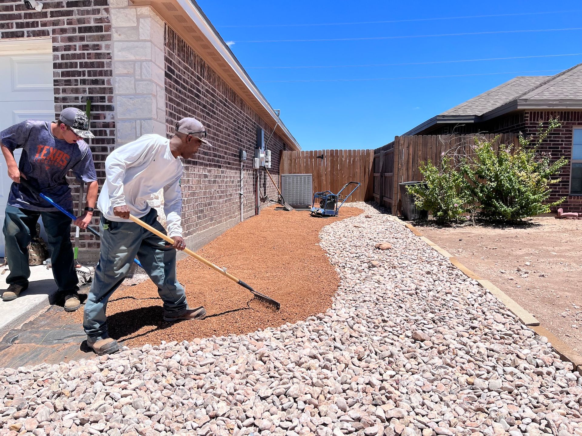 Two men raking gravel in a residential yard. One rakes brown gravel, the other rakes white. Sunny day.