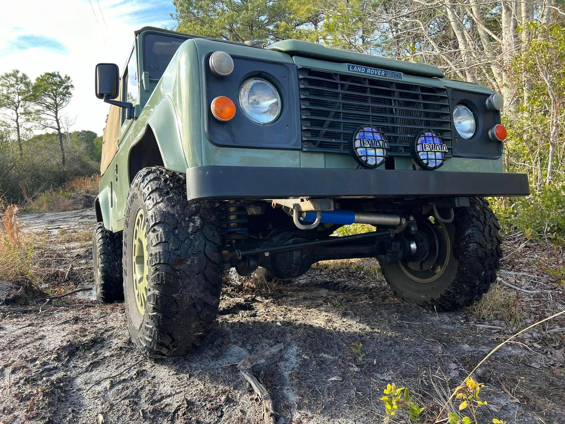 Green Land Rover Defender, modified for off-roading, parked on a sandy path in a wooded area.