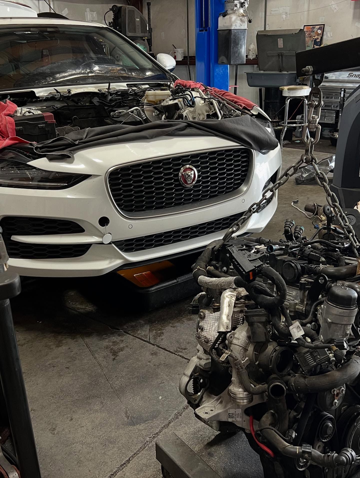 White Jaguar car with open hood at a repair shop, engine removed and ready for replacement.